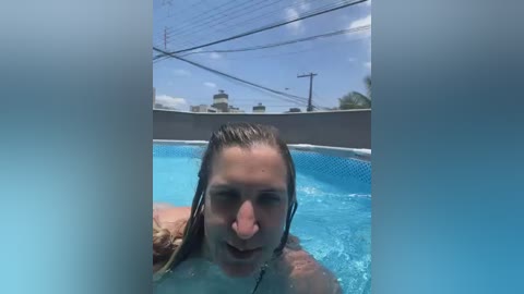 Video of a young woman with wet, blonde hair and a large nose ring, swimming in a blue, tiled pool under a bright blue sky.