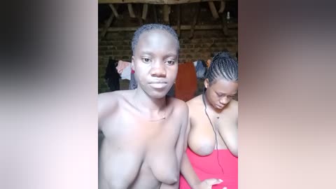 Video of two African women, one topless with small breasts, the other in a red top, standing in a rustic brick-walled room with wooden beams.