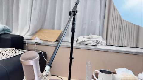 Video of a minimalist, cluttered room with a white bed, beige walls, a window with sheer curtains, a wooden table holding a microphone stand, a glass, and a white mug.