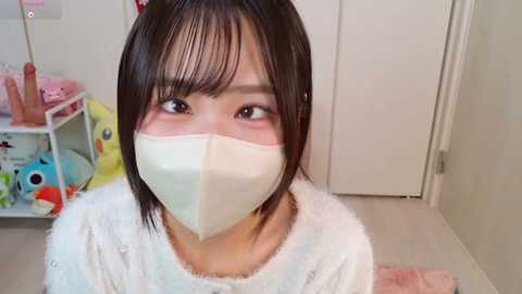 A video of an Asian woman with short black hair, wearing a white fluffy sweater and a white surgical mask, standing in a cluttered, pastel-colored room with toys and a stuffed animal.