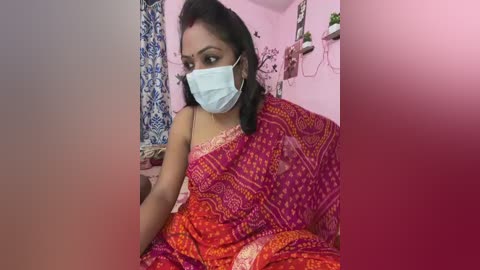 Media: A video of a woman in a pink saree with intricate designs, wearing a blue surgical mask and sitting in a pink-painted room with white shelves and decorative items.