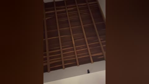 A video of a rustic, wooden ceiling with horizontal beams and vertical supports, set against a dark, shadowy background. The ceiling appears to be part of a traditional, possibly African or South American, building.