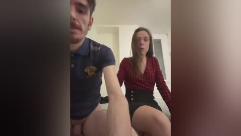 Video of a man and woman in a dimly lit room. The man, in a navy polo, sits on the bed, looking serious. The woman, in a red checkered top and black skirt, sits on the floor, gazing up.