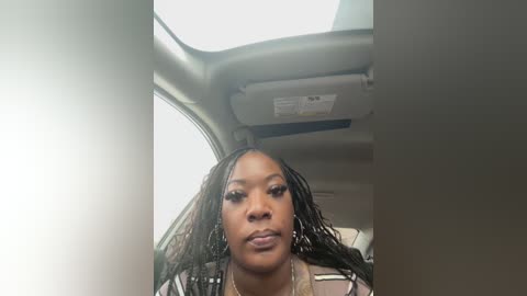 Media: Video of a Black woman with braided hair and light makeup inside a car, wearing a white top.