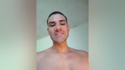 Video of a young, shirtless man with short dark hair and a light skin tone, wearing a neutral expression. He stands against a plain, off-white wall in a minimalist setting.