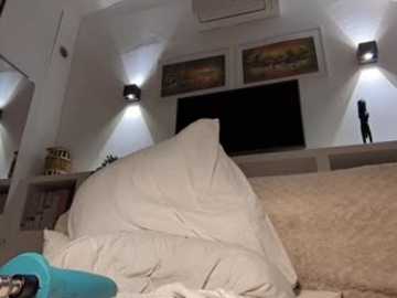 Video of a dimly lit, cozy bedroom with beige bedding, two mounted wall lights, framed artwork, a beige cushion, and a blue object on the bed.