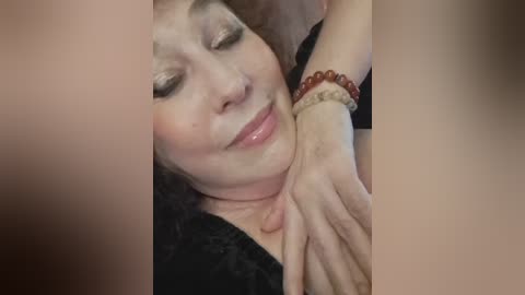 Video of a woman with light skin, closed eyes, and a serene expression, lying on a bed. She wears a black robe and a red beaded bracelet.