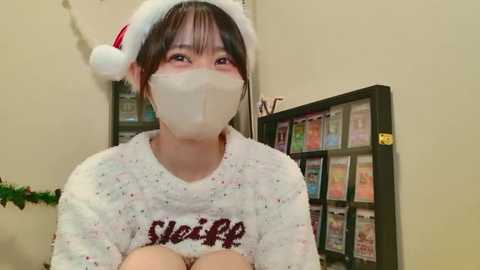 Video of an Asian woman wearing a festive Santa hat, white mask, and \"Weir\" sweater, standing in a cozy room with holiday decorations and a bookshelf.