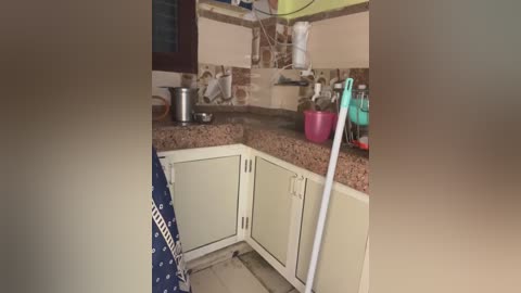 Video of a small, cluttered kitchen with beige cabinets, brown granite countertops, a blue apron hanging, a white broom leaning, and various kitchen utensils and appliances.