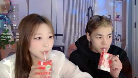 Video of two young East Asian women drinking from red-and-white milk cartons, one with long hair, the other with short hair. Background shows a modern, brightly lit room with shelves and a wall calendar.