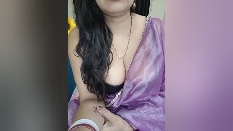 Media: Video of a dark-skinned woman with long, wavy black hair, wearing a purple sari with a black bra visible, sitting indoors.