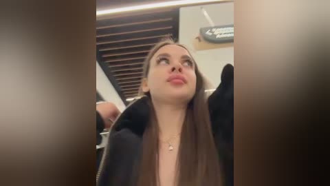 A video of a young woman with long brown hair, wearing a black jacket, sitting in a modern interior with dark wooden slats and a beige wall.