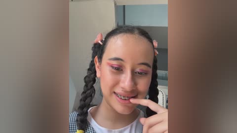 Video of a young woman with dark skin, braided hair, and pink ribbons, wearing a plaid shirt, smiling and showing braces, taken indoors with a blurred background.