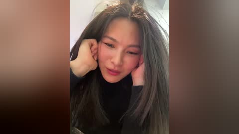 Video of a young Asian woman with long, straight black hair, wearing a black turtleneck, holding her head with both hands. Background is blurred with a warm, brown gradient.