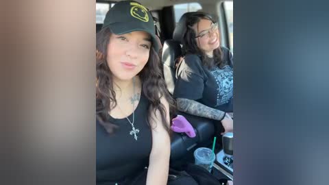 Video of two young women with long, dark hair, wearing black tank tops, sitting in a car. One wears a black cap with a yellow smiley face.