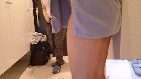 Video of a woman in a blue dress and gray sneakers bending over in a bedroom with beige walls, a black suitcase, and a white bed.