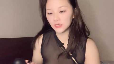Video of an East Asian woman with long, straight black hair, wearing a sheer black top, standing indoors against a neutral background.