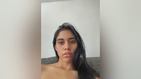 Video of a young woman with long black hair, light brown skin, and a slim physique, topless, sitting on a gray couch. The background is a plain white wall.