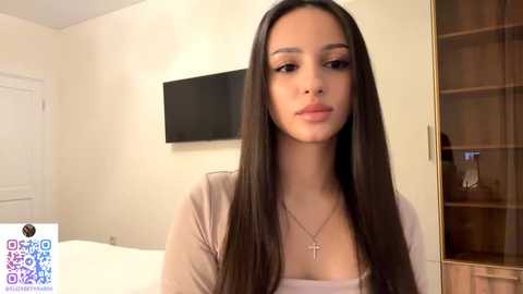 Video of a young woman with long, straight, dark brown hair and light skin, wearing a beige top, standing in a modern, beige-walled bedroom with a flat-screen TV and wooden closet in the background.