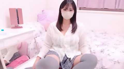 Video of an Asian woman with long black hair, wearing a white face mask, white cardigan, and grey skirt, sitting on a bed with floral bedding, in a softly lit room.