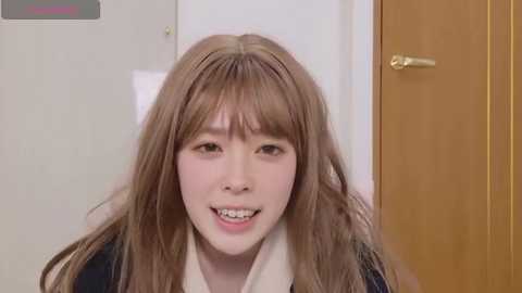 Media: Video of a young Asian woman with long, straight brown hair and braces, smiling, wearing a black jacket over a white shirt, indoors near a wooden door.