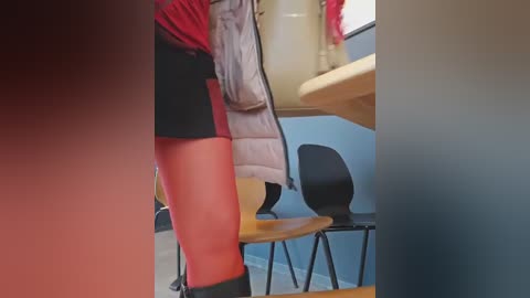Video of a person in a classroom, wearing a red jacket, black shorts, and black boots, leaning over a desk with a beige chair. Background features a blue wall and another chair.