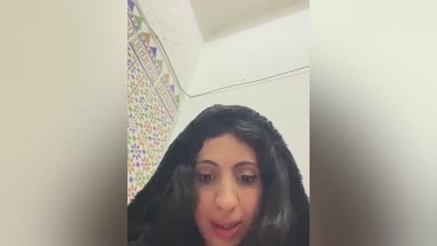 Video of a young woman with long, dark hair and olive skin, standing in a tiled bathroom with colorful mosaic patterns on the wall, making a funny face.