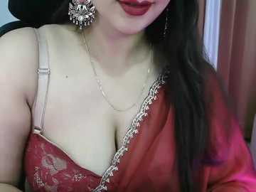Video of a fair-skinned woman with long black hair, wearing a red saree with intricate silver embroidery, adorned with large silver earrings. She has full red lips. Background shows blurred indoor setting with soft lighting.