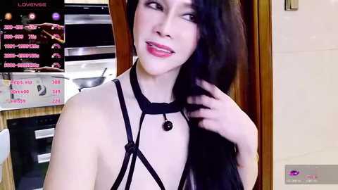 Video of a pale, slender woman with long black hair, wearing a black strappy harness top, smiling in a modern kitchen.