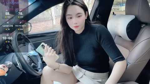Media: Video of an East Asian woman with long black hair, wearing a black turtleneck and beige skirt, sitting in a modern car, using a smartphone, displaying virtual reality game stats.