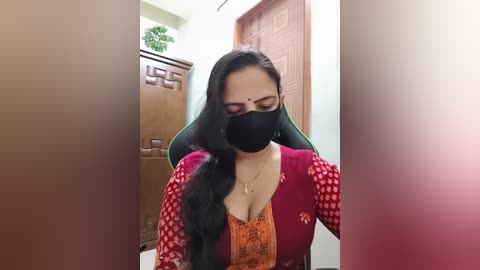 Video of a South Asian woman with long black hair, wearing a red and orange floral-patterned blouse, black mask, and a green chair, indoors, wooden furniture in background.