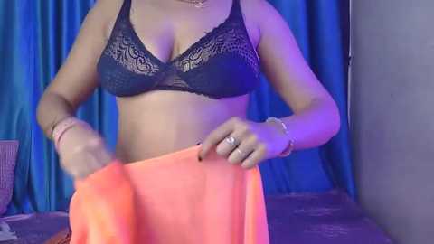 Video of a woman with medium skin tone, wearing a black lace bra and orange cloth, standing in front of a blue curtain.