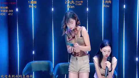 Video of two Asian women in casual, beige outfits on a stage with blue, lit curtains in the background.
