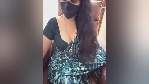 Video of a South Asian woman with medium skin tone, wearing a black mask, black top, and blue patterned sari, seated indoors.