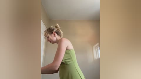 Video of a slender, fair-skinned woman with blonde hair in a bun, wearing a strapless green dress, standing in a beige-walled room, partially obscured by a white door.