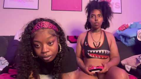 Video of two young Black women in a cozy bedroom, one playing a video game, wearing a black crop top, the other with curly hair, wearing a black crop top.