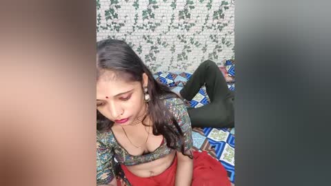 A video of a young woman with long black hair, wearing a colorful top, red pants, and a bindi, lying on a patterned bedspread in a room with a white brick wall and green ivy.