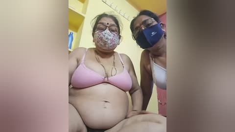 Video of two women wearing face masks, one in a pink bra and floral face mask, the other in a blue bra, standing in a yellow-walled room with a red door.