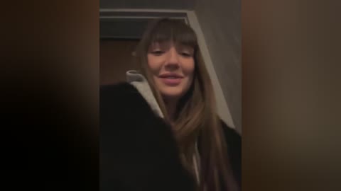 A blurry video of a young woman with long, straight, brown hair, wearing a black jacket and white shirt, smiling softly in a dimly lit, narrow hallway.