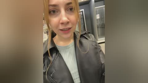 Video of a young woman with fair skin and blonde hair, wearing a black leather jacket over a light blue sweater, standing indoors with a blurred background.