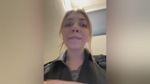 Video of a young, fair-skinned woman with light brown hair, wearing a black jacket over a grey lace top, standing in a dimly lit, beige-walled hallway.