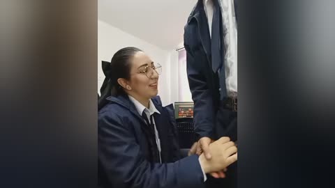 Video of a woman in glasses and a navy jacket, kneeling in front of a standing man in a suit. They are in a dimly lit office with a desk and computer monitor in the background.