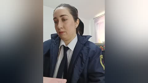 Video of a female police officer with medium-length dark hair, wearing a navy blue uniform with a yellow badge, a white shirt, and a black tie, seated indoors, holding a pink phone.