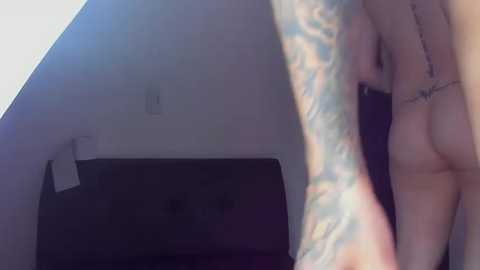 Video of a partially nude woman with light skin, tattoos on her arm, standing in a dimly lit room with a white wall and a dark floor.