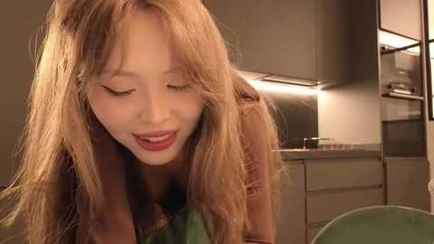 A video of a young Asian woman with long, wavy, light brown hair, wearing a green top, smiling softly. She is in a modern kitchen with dark cabinets, a countertop, and a stainless steel oven.
