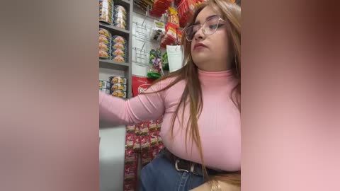 A video of a young woman with long blonde hair and glasses, wearing a pink turtleneck and blue jeans, reaching into a red and white snack aisle in a supermarket.