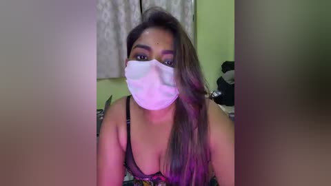 Video of a young South Asian woman with long, straight brown hair, wearing a black bra and a white surgical mask, set against a dimly lit, cluttered bedroom background.