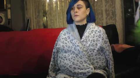 Video of a fair-skinned person with blue hair, wearing a leopard print blanket, sitting on a red couch in a dimly lit room with patterned curtains.