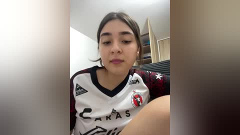 Video of a young woman with light brown skin and long dark hair, wearing a red and white soccer jersey with \"Carranza\" and \"Soccer\" logos, sitting on a bed in a dimly lit room.