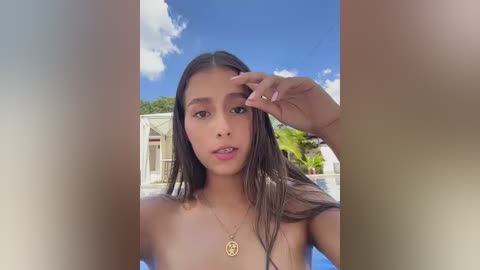 A video of a young woman with long dark hair, tanned skin, and a gold necklace, standing outdoors by a pool, with a blurred background of a house and trees.